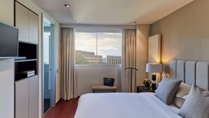 A modern hotel room with a large bed, a TV, a bench, a nightstand with a lamp, and a view of the city through the window. - Hyatt Regency Madrid Residences (Madrid)
