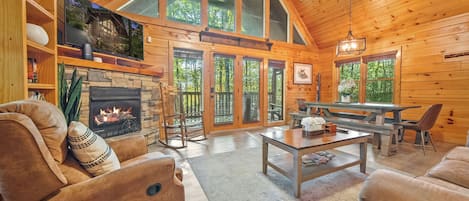 Un interior de cabaña espacioso con chimenea de piedra, muebles de madera y ventanas amplias que dan a un bosque.