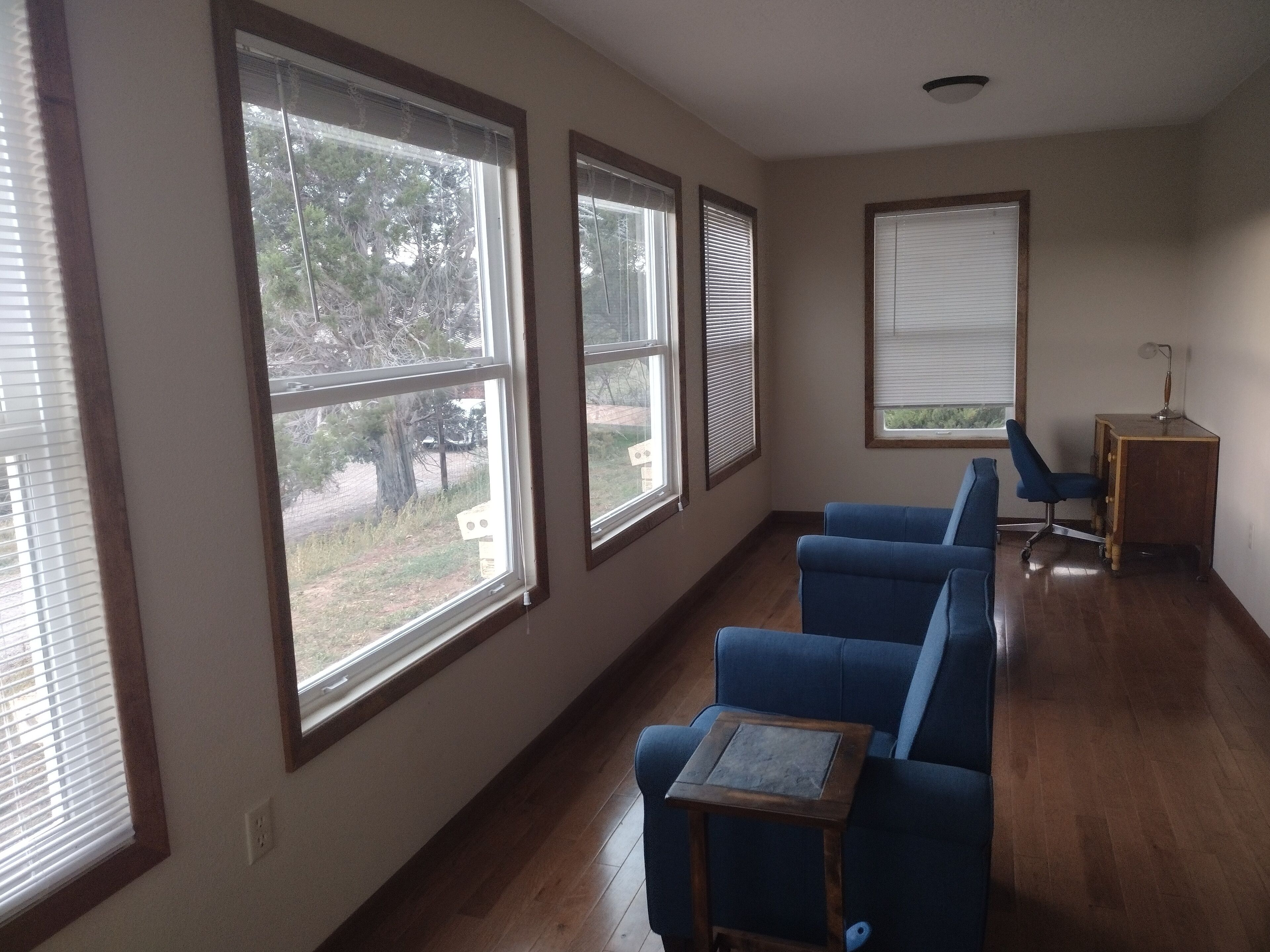 Una habitación con ventanas amplias, un sofá azul, una mesita de madera y un escritorio con una lámpara.
