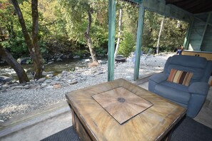 A wooden table with a metal center, a blue sofa with a striped pillow, and a view of a river with rocks and trees. - ❤Riverfront Paradise - Butterfly Ranch on the River❤ (Dunsmuir)