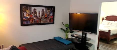 A modern living room with a flat-screen TV, a cityscape painting, a sofa, a coffee table, and a bed in the background.