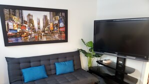 Ein Wohnzimmer mit grauem Sofa, zwei blauen Kissen, einem großen Stadtbild in einem Bilderrahmen und einem Sony-Fernseher auf einem Standfuß.