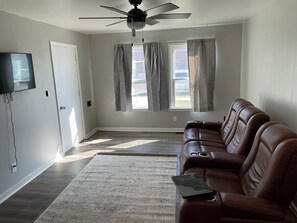 A living room with a brown leather sofa, a flat-screen TV mounted on the wall, a ceiling fan, and a window with curtains.