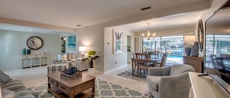 A spacious living room with a sofa, a coffee table, and a TV. There's a dining area with a table and chairs, and a kitchen in the background.