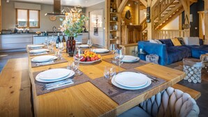 Un coin repas avec une longue table en bois dressée pour le repas, une cuisine en arrière-plan et un escalier menant à un niveau supérieur.