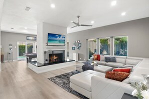 A spacious living room with a fireplace, a large TV, and a ceiling fan. - Spacious Home with Heated Pool & Billiard sleep 18 (Dallas)