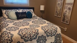 A bedroom with a bed, a window with blinds, a bedside lamp, and two wall-mounted pictures.