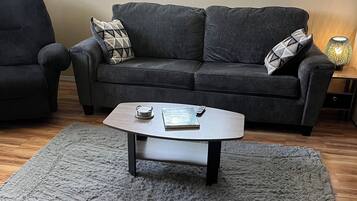A living room with a grey sofa, a grey armchair, a round coffee table with a book and a remote control, a grey shaggy rug, wooden flooring, a window with blinds, and a wall-mounted picture.