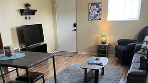 A living room with a wooden floor, a gray rug, a dining table with chairs, a sofa, a TV, and a small table with a lamp.