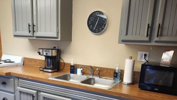A kitchen with grey cabinets, a wooden countertop, a coffee maker, a sink, a clock, and a microwave.