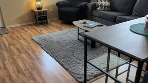 A modern living room with a wooden floor, a grey rug, a black armchair, a grey sofa, and a small side table with a lamp.
