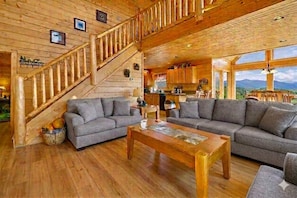 A wooden interior with a staircase, grey sofas, a wooden coffee table, and a kitchen area.