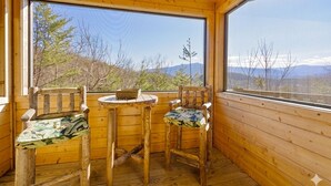 Une cabane en bois avec deux chaises et une petite table, de grandes fenêtres offrant une vue sur les arbres et les montagnes.