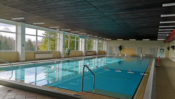 An indoor swimming pool with clear glass walls, a tiled floor, and a ceiling with linear lighting fixtures.
