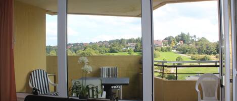A balcony with a view of a green landscape and houses. There is a table with chairs and a potted plant.