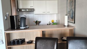 A modern kitchen with white cabinets, a stainless steel range hood, and three pendant lights. There are two black chairs at a wooden table, and a coffee maker on the counter.