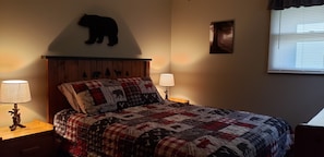 A bedroom with a wooden headboard, a quilted bedspread with animal prints, two bedside lamps, a wall-mounted bear decoration, and a framed picture.