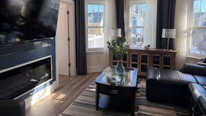 A modern living room with a flat-screen TV, a fireplace, a coffee table, a sofa, and large windows with curtains. - Renovated 2nd-Floor Home (Boston)