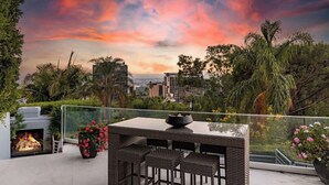 A balcony with a glass railing, a fire pit, wicker furniture, and a view of the cityscape. - Villa Lareira (Los Angeles)