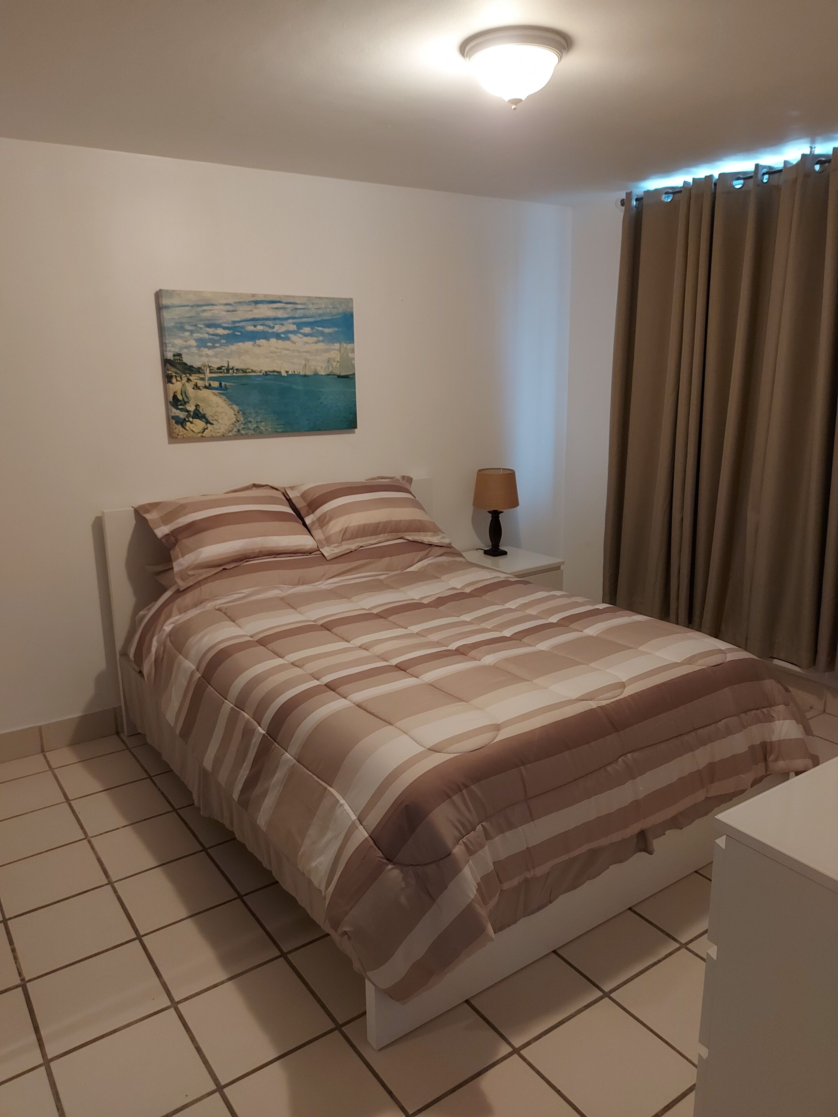 Una cama bien tendida con ropa de cama a rayas, una lámpara de noche y un cuadro con una escena costera en la pared.
