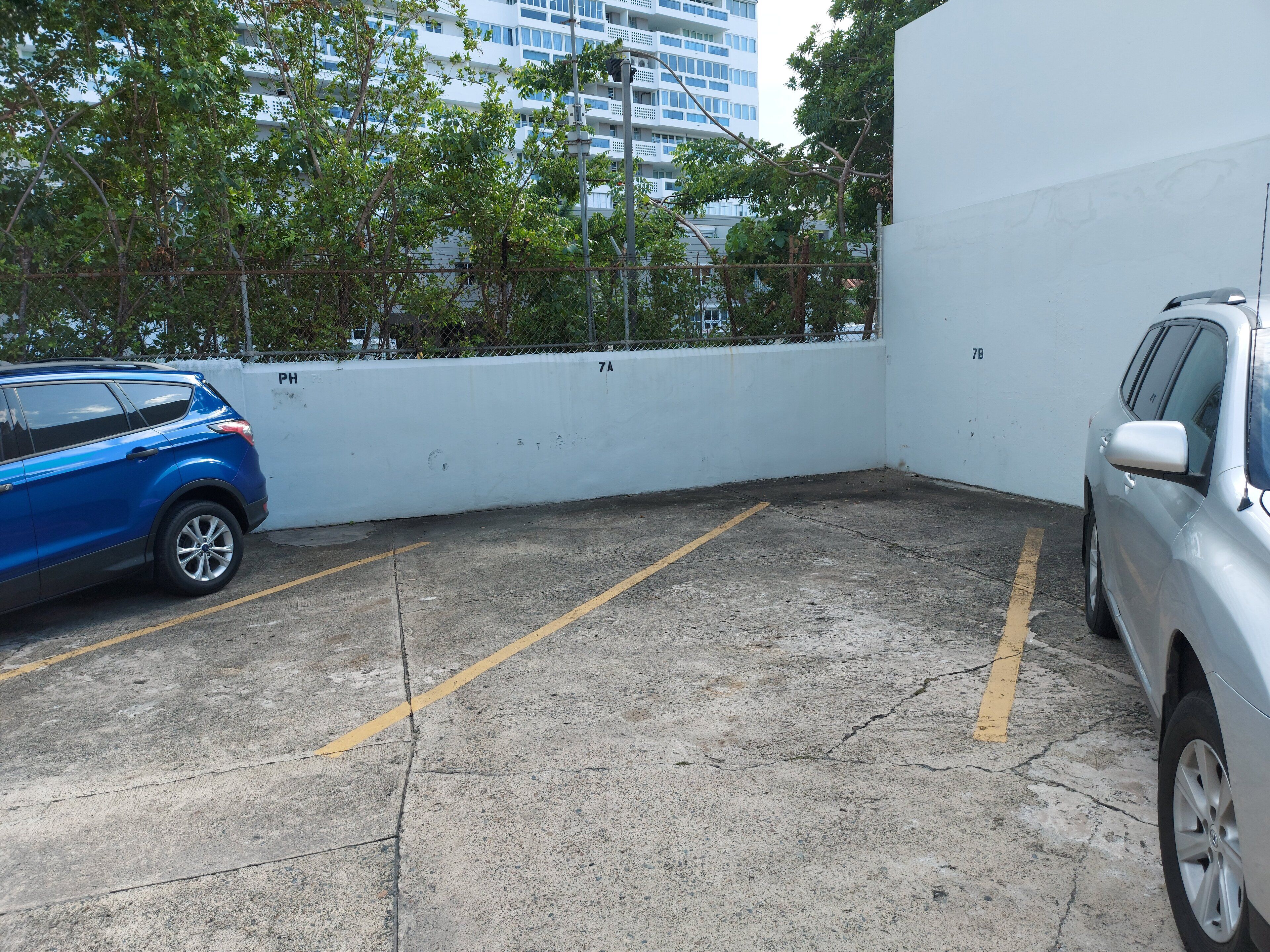A parking area with two cars, a white wall, and a building in the background.