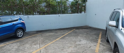 A parking area with two cars, a white wall, and a building in the background.