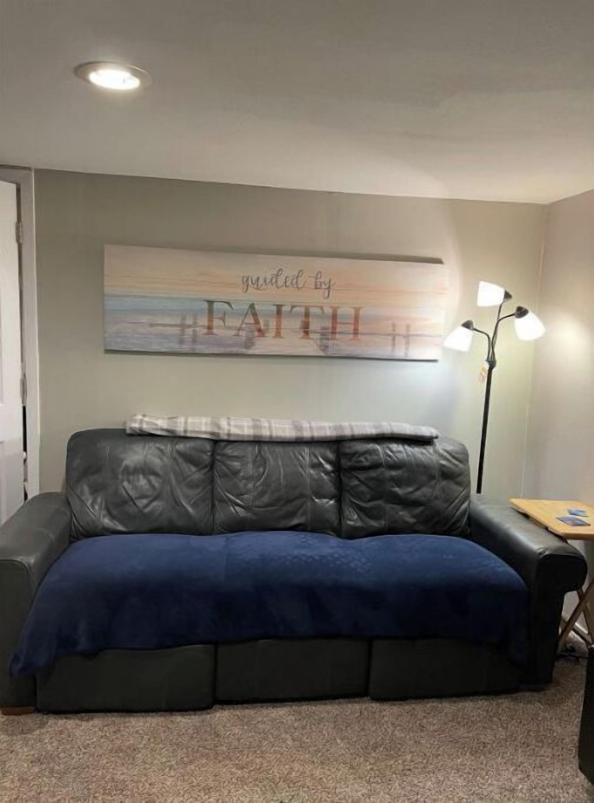 A room with a dark leather couch, a blue throw blanket, a wall-mounted artwork with the text "guided by FAITH", and a floor lamp.