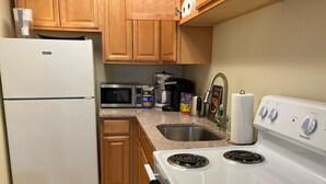 Una cocina con refrigerador blanco, microondas, estufa y pileta. Hay alacenas de madera, una cafetera y diversos objetos de cocina sobre la encimera.
