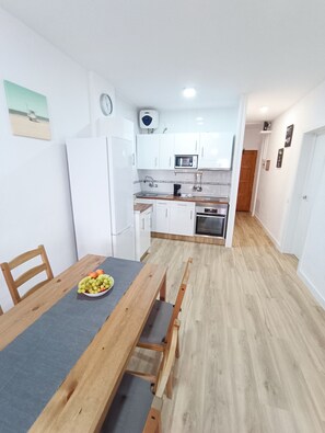 A modern kitchen with white cabinets, a microwave, and a coffee maker. There is a dining table with a bowl of fruit and a wooden bench.