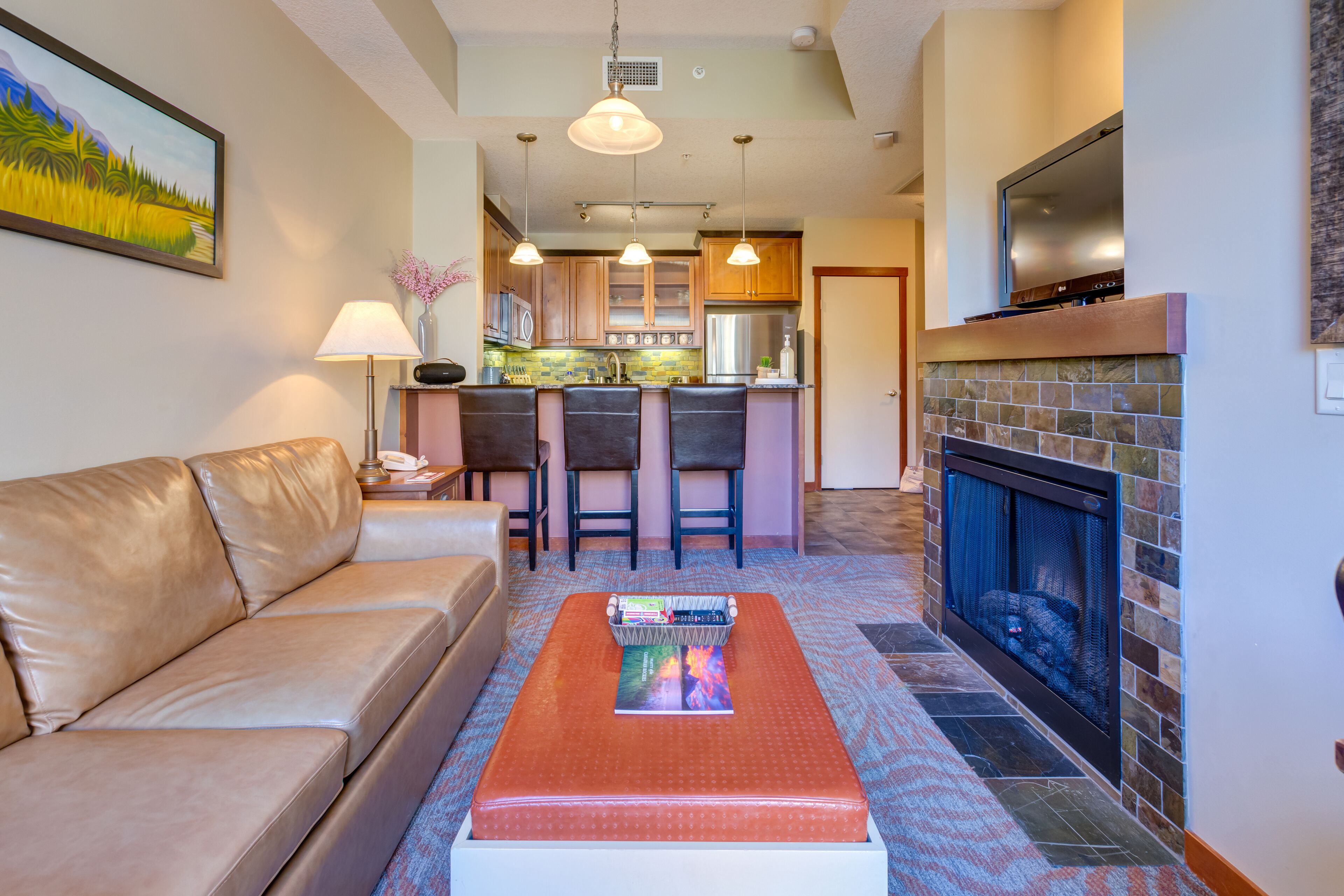 A living room with a sofa, a coffee table with books, a fireplace, and a kitchen area in the background.