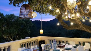 An outdoor dining area with a table set for four, surrounded by white chairs, under hanging lights and a tree.