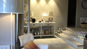 A modern living room with a white leather sofa, a matching ottoman, and a wooden coffee table. A white lamp and a bookshelf with decorative items are also visible.