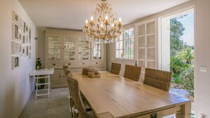 A dining area with a wooden table and wicker chairs, a chandelier, and a view of greenery outside.