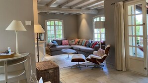 A cozy living room with a sectional sofa, a small round table, and a wicker ottoman. There are two windows with curtains, and the room has exposed wooden beams on the ceiling.