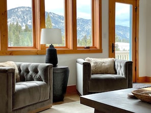 A room with two leather sofas, a wooden coffee table, and a lamp. Large windows with wooden frames offer a view of a mountainous landscape. - Spectacular Private Big Sky 5 Bedroom Home - BEST LOCATION ! (Big Sky)