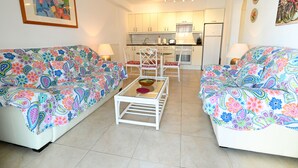 A living room with two colorful sofas, a glass coffee table, and a kitchen in the background. - A. Parque Santiago 1-1 bed-pool-sea front location. Sunny. (Playa de la Américas)