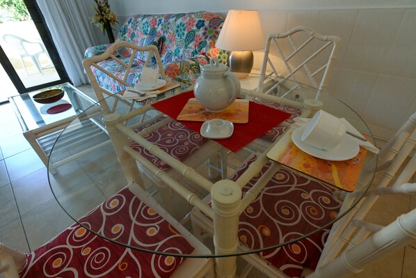 A dining area with a glass table, red patterned placemats, white chairs, and a floral sofa.