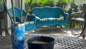 Un espace patio aménagé avec un canapé en osier bleu, une table en métal avec un seau noir et une plante ornementale de type « oiseau de paradis » dans un vase bleu.