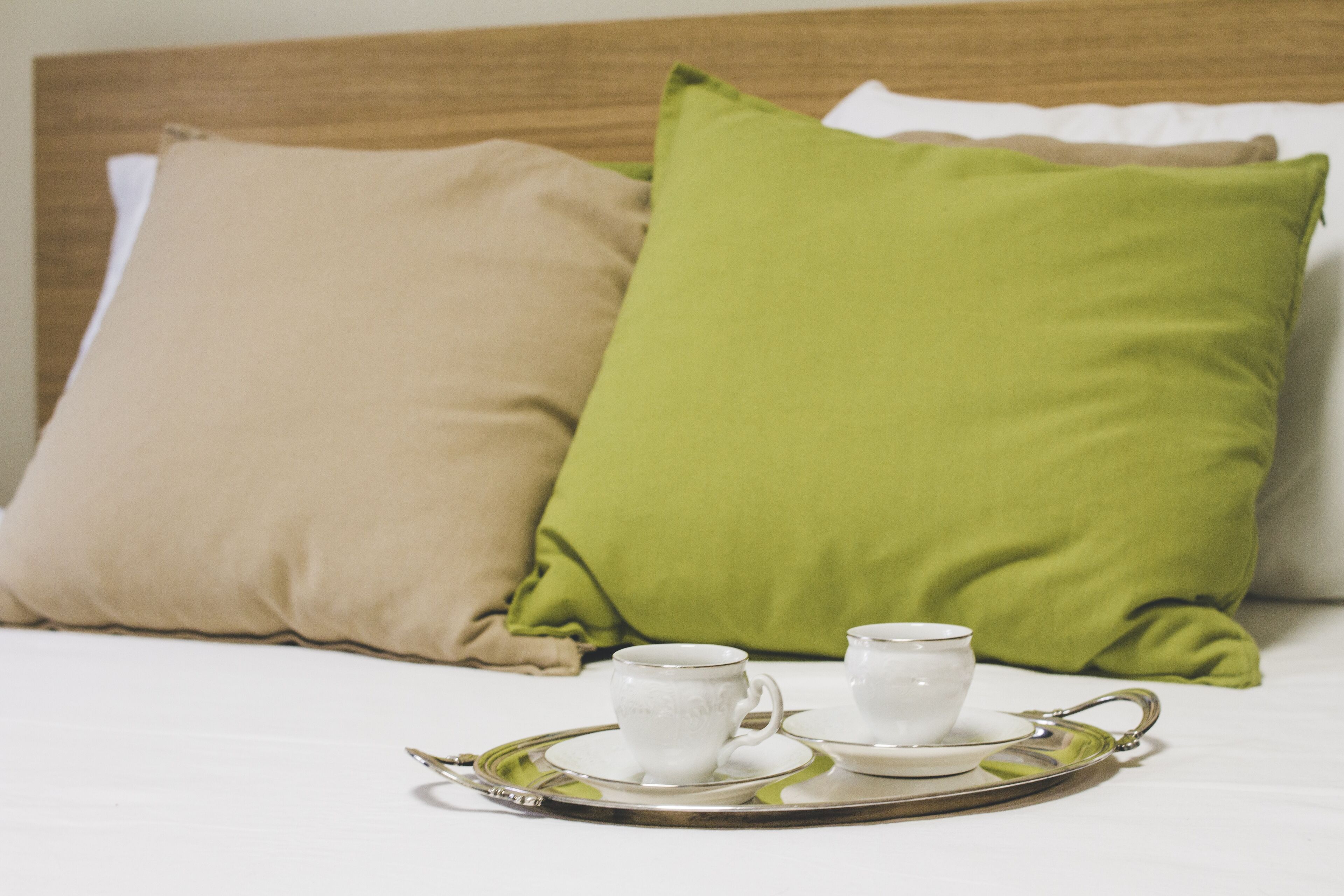 Een netjes opgemaakt bed met twee kussens, één beige en één groen, en een dienblad met twee witte kopjes en schoteltjes.