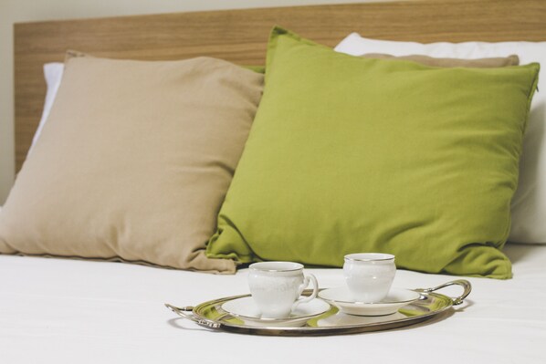 Una cama bien hecha con dos almohadas, una beige y otra verde, y una bandeja con dos tazas y platillos blancos.