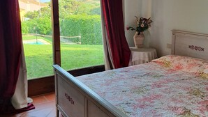 Quarto com cama, janela com cortinas, uma mesinha com um vaso e vista para uma paisagem verde.