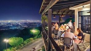 Un área de comedor al aire libre con mesas y sillas, con vista a un paisaje urbano nocturno.