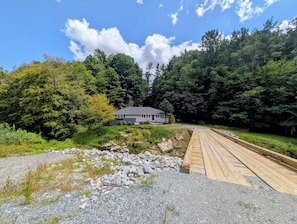 Una casa con porche, rodeada de un bosque denso, y un puente de madera que la conecta.