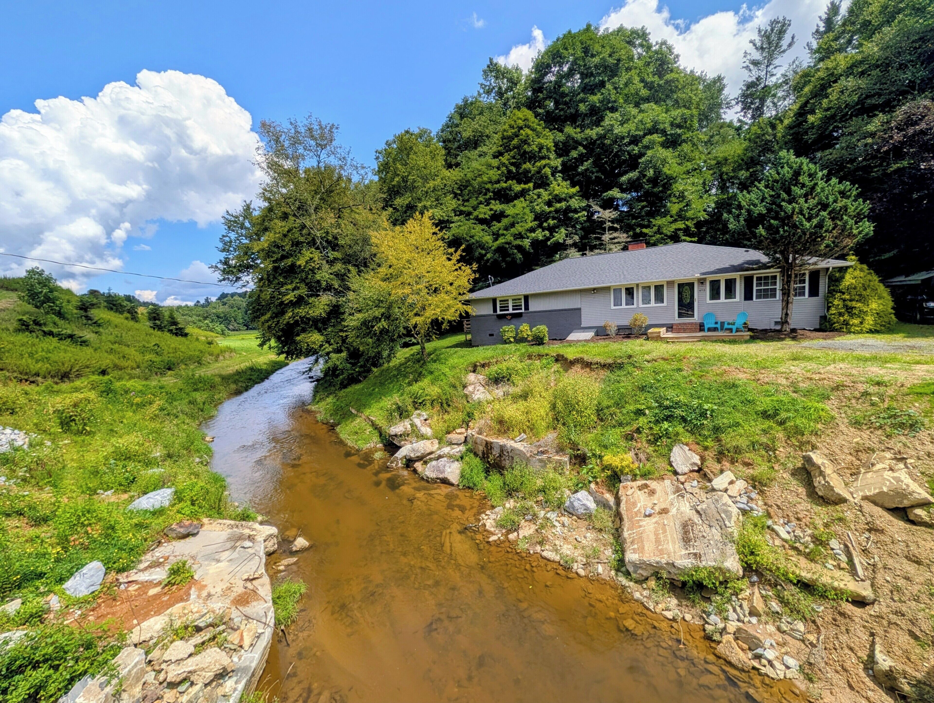 A house with a front porch, a small stream, and surrounding greenery.