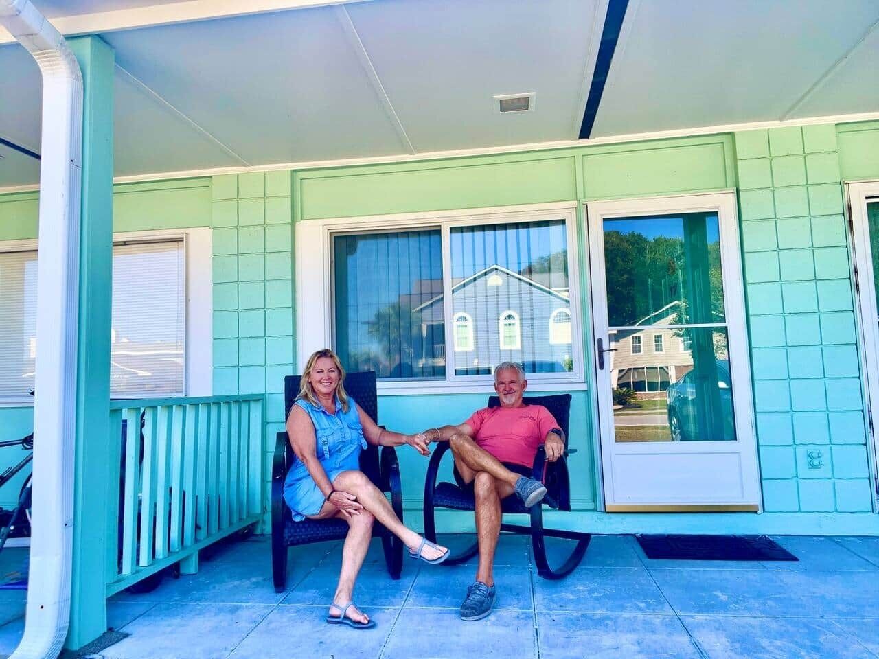 Eine Veranda mit zwei sitzenden Personen, einer Glastür und einem Blick auf ein Haus im Hintergrund.