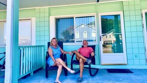 A porch with two people seated on chairs, a door with a glass panel, and a view of a house in the background. - Love Shak romantic beach condo , pets ok (North Myrtle Beach)