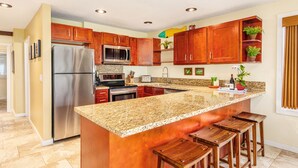 A kitchen with wooden cabinets, a stainless steel refrigerator, a granite countertop, and bar stools. - Ocean Front, Ground Level 1 Bedroom with King, Sleeps 4, with Cold AC (Kapaʻa)
