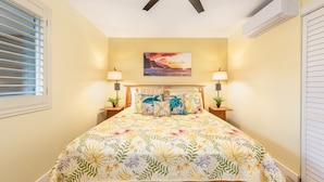 A bedroom with a bed, two bedside tables, a nightstand, a ceiling fan, a wall-mounted air conditioner, and a window with blinds. - Ocean Front, Ground Level 1 Bedroom with King, Sleeps 4, with Cold AC (Kapaʻa)