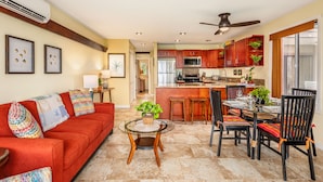A spacious living room with a red sofa, a glass coffee table, and a dining area with a table set for four. - Ocean Front, Ground Level 1 Bedroom with King, Sleeps 4, with Cold AC (Kapaʻa)