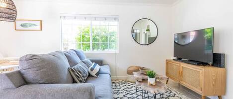 Una sala de estar moderna con un sofá gris, un mueble de televisión de madera, un ventilador de techo y un ventanal con vistas a zonas verdes.